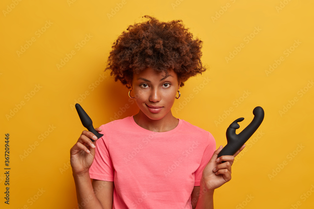Isolated shot of African American woman poses with sex toys, uses butt plug for anal sex and rabbit vibrator to release sexual tension and boost your mood, increases sexual function gets self pleasure Isolated shot of African American woman poses with sex toys, uses butt plug for anal sex and rabbit vibrator to release sexual tension and boost your mood, increases sexual function gets self pleasure