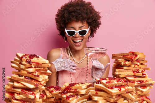 Joyful elegant woman expresses positive emotions and has fun at party  stands near table with stack of delicious bread toasts  holds glass of cocktail  wears sunglasses  dress and lace gloves