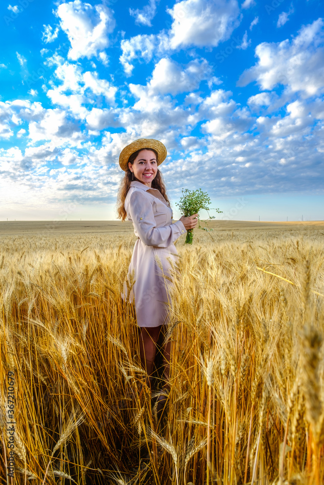Smiling young long-haired woman in straw hat smiling holds a wild flower bouquet in wheat field at dawn. Model looks direct at camera, vertical orientation Smiling young long-haired woman in straw hat smiling holds a wild flower bouquet in wheat field at dawn. Model looks direct at camera, vertical orientation
