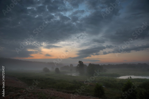Foggy sunrise over a meadow and a river with dark clouds in the sky.