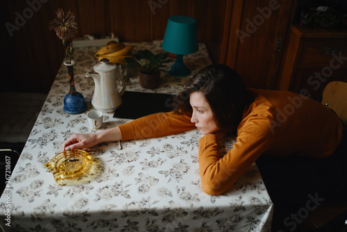 A sad girl in a brown sweater sits in the kitchen with her head resting on her hands on the table