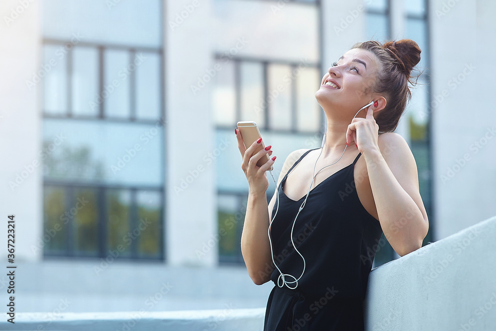 Fototapeta premium beautiful girl poses in the city, holding a smartphone and listening to music with headphones