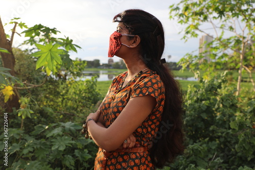 A young Indian Mexican Lady  girl against the sun standing with home made Corona protection face Mask in colourful dress with eye makeup and posing talking on the phone photo