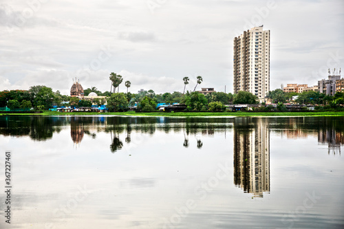 Landscape Wide angle shot of the buildings temple and colony with the reflection in the rain water pond and green coconut tree boys fishing with stick in Mumbai Maharashtra India on 27 July 2020