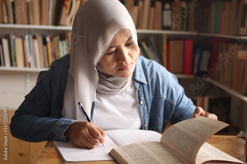 Asian muslim woman studying in library, exam preparation concept. Female college student doing research and making notes in her book