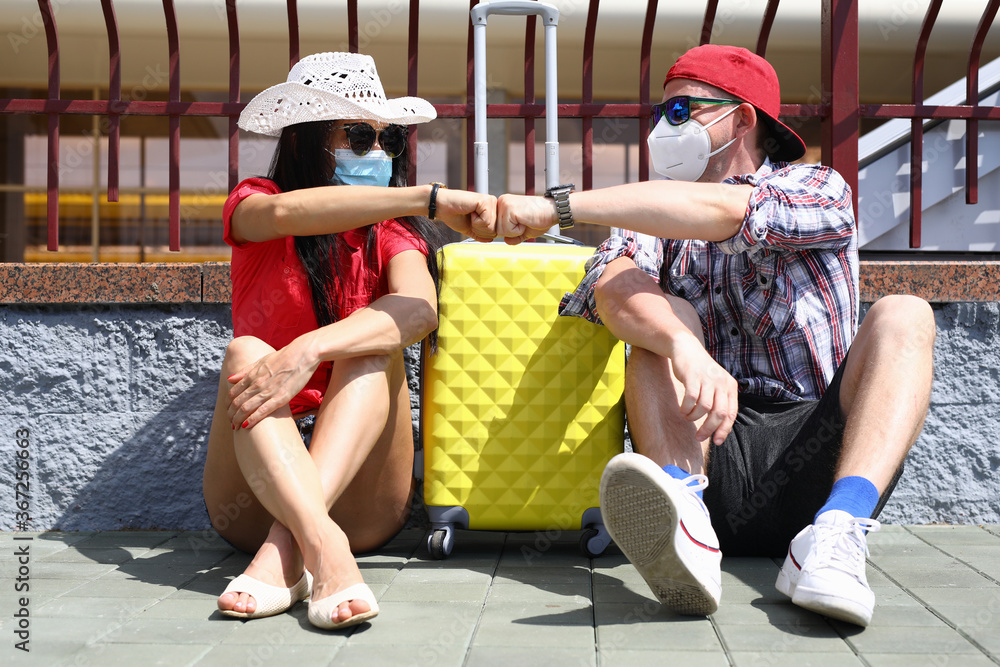 Handsome man and beautiful woman sit on pavement on platform of train station with yellow suitcase in protective mask fist to fist. T Couple wait for departure of train with luggage. - obrazy, fototapety, plakaty 