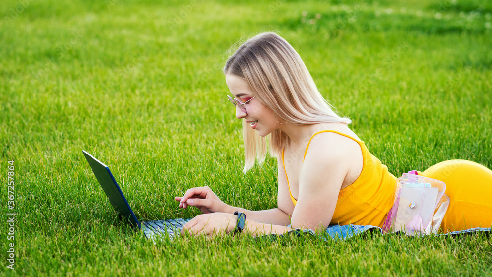 Girl freelancer. Attractive woman using laptop lying on lawn in park surfing the net or preparing for exams. Technology, education and remote work concept. Freelance Worker. Girl freelancer. Attractive woman using laptop lying on lawn in park surfing the net or preparing for exams. Technology, education and remote work concept. Freelance Worker.