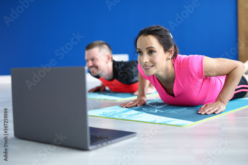 Couple look into laptop on floor. Woman and man do push-up on gymnastic mat and watch video in loptop. Drawing up a program for sport at home. Remote exercises online. photo