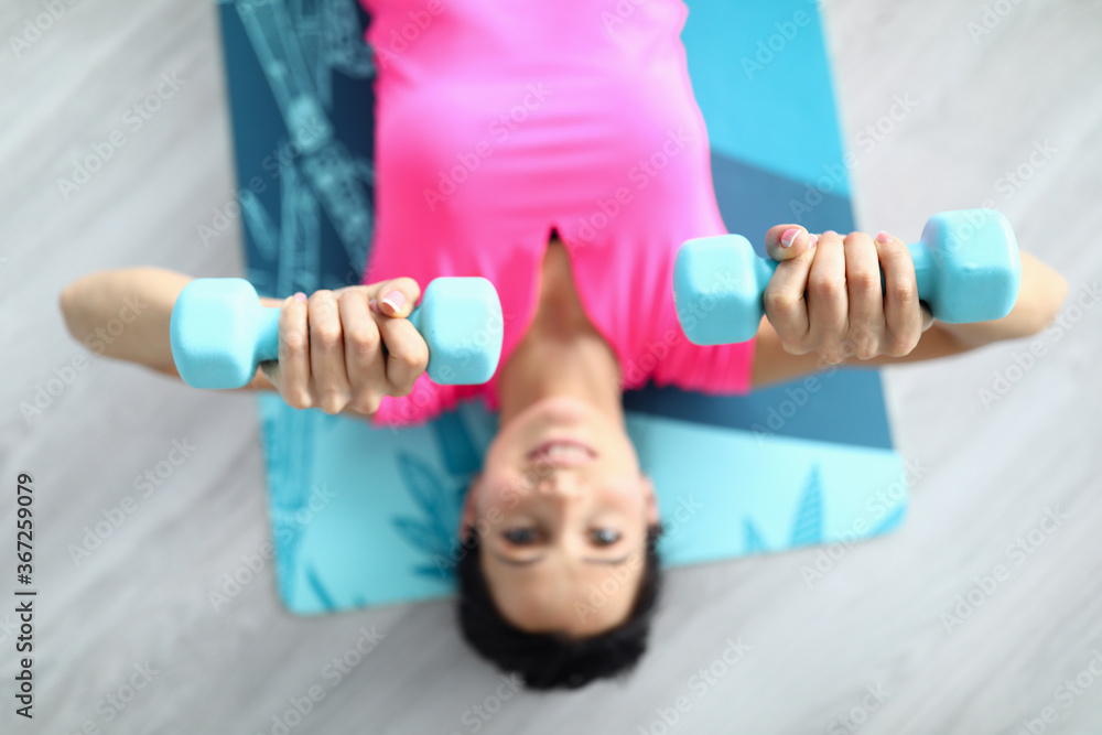 Beautiful woman in pink T-shirt lie on gymnastic mat and rais her arms with dumbbells up above herself. Trainer use sports equipment to exercise and build muscle. Professional nutrition and weight Beautiful woman in pink T-shirt lie on gymnastic mat and rais her arms with dumbbells up above herself. Trainer use sports equipment to exercise and build muscle. Professional nutrition and weight