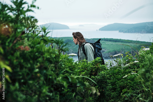 close up young long-haired man with a backpack and a photo tripod walking along a trail through the grass at the edge of a cliff with sky and sea background.. Travel and outdoor concept.