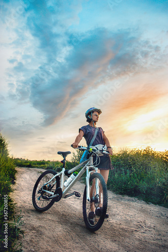 Young woman and mountain bike stands in the rural green field at summer evening. Adventure summer leisure time.