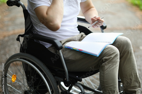 Man sit in wheelchair and take note in notebook in park. Disabled person keep diary and hold pen in his hand and notebook on his lap. Preparation of classes for rehabilitation after surgery.