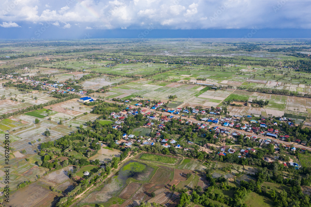A top down aerial view of a small country town with traditional houses with orange roofs, a red dirt road, rice fields, and palm trees in the jungle in Cambodia.