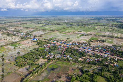 A top down aerial view of a small country town with traditional houses with orange roofs, a red dirt road, rice fields, and palm trees in the jungle in Cambodia. © Nhut
