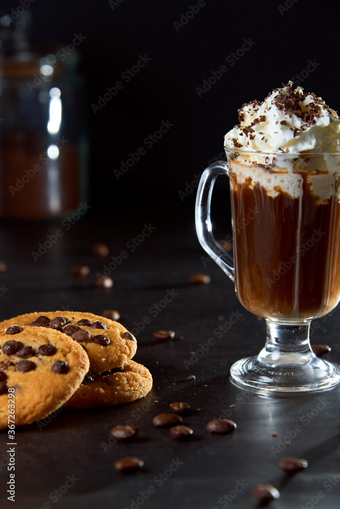 Close up of a glass cup of coffee with whipped cream and chocolate on it, chocolate chip cookies and roasted coffee beans on dark background. Concept of ready to eat food, tasty snack. Selective focus Close up of a glass cup of coffee with whipped cream and chocolate on it, chocolate chip cookies and roasted coffee beans on dark background. Concept of ready to eat food, tasty snack. Selective focus