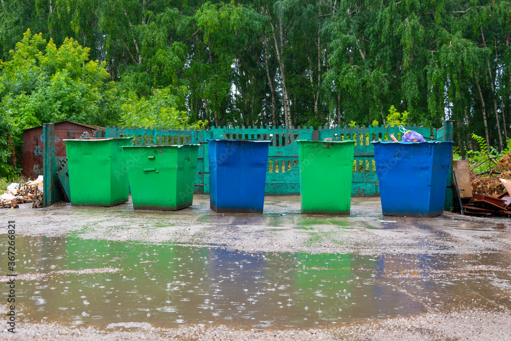 An area with multi-colored waste bins flooded with pouring rain. Puddles of water