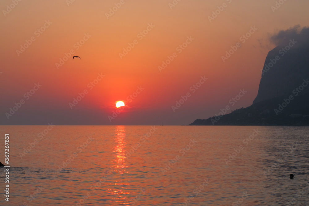 Seascape sunset on the sea, cloud on the top of the mountain, the sun sets in the clouds on the horizon, the sea and sky are orange-pink