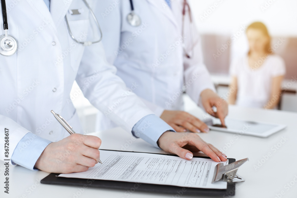 Unknown woman-doctors at work with patient at the background. Female physicians filling up medical documents or prescription while standing in sunny hospital reception desk, close-up. Health care Unknown woman-doctors at work with patient at the background. Female physicians filling up medical documents or prescription while standing in sunny hospital reception desk, close-up. Health care