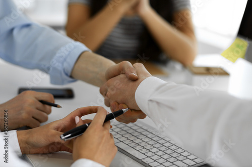 Two businessmen are shaking hands in office while sitting at the desk, close-up. Colleagues applauding of success meeting end. Business people concept