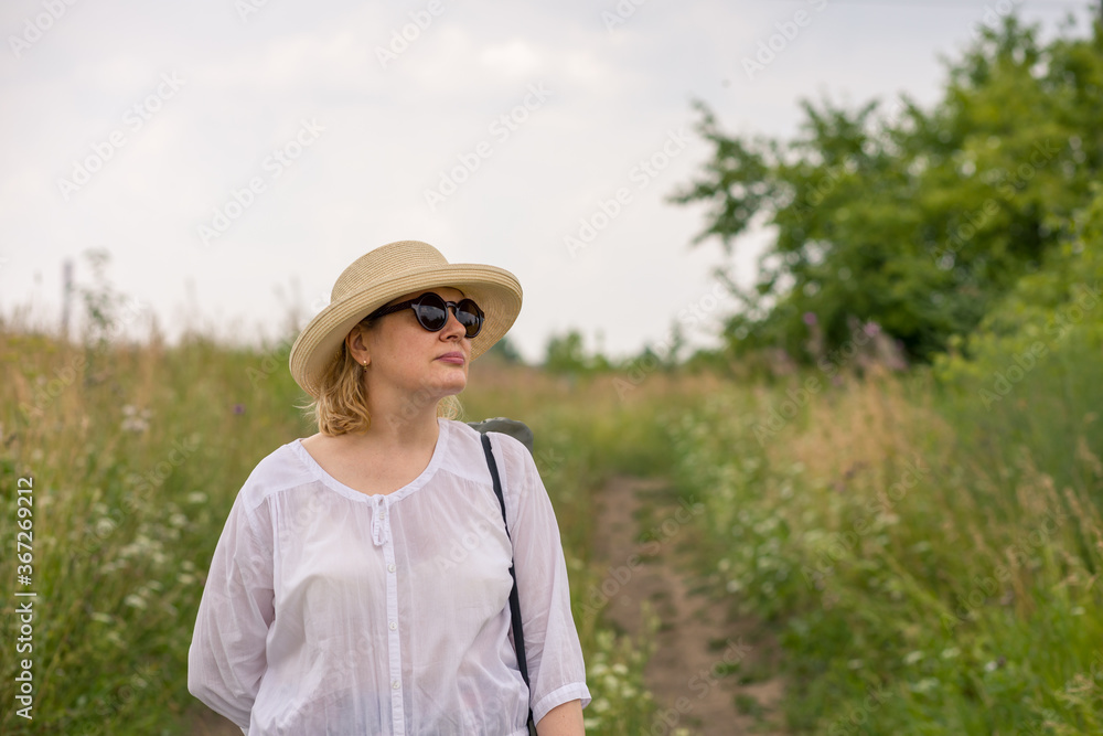 An elderly woman in an elegant straw hat walks along a rural road, enjoying the scenery. The concept of an active lifestyle for older people.