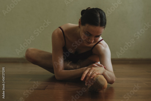 Beautiful graceful young ballerina in pointe shoes sitting on the brown wooden floor. Ballet practice and stretching. Female stretching indoors toes professional arts. Female fit and healthy lifestyle