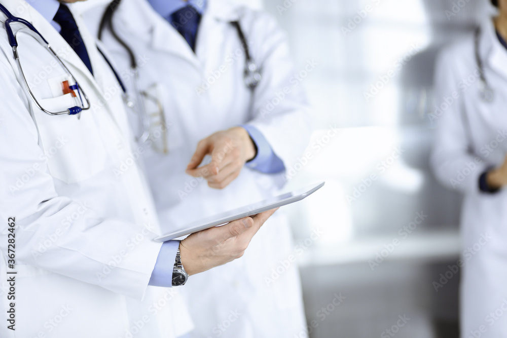 Group of unknown doctors use a computer tablet to check up some medical names records, while standing in a hospital office. Physicians ready to examine and help patients. Medical help, insurance in Group of unknown doctors use a computer tablet to check up some medical names records, while standing in a hospital office. Physicians ready to examine and help patients. Medical help, insurance in