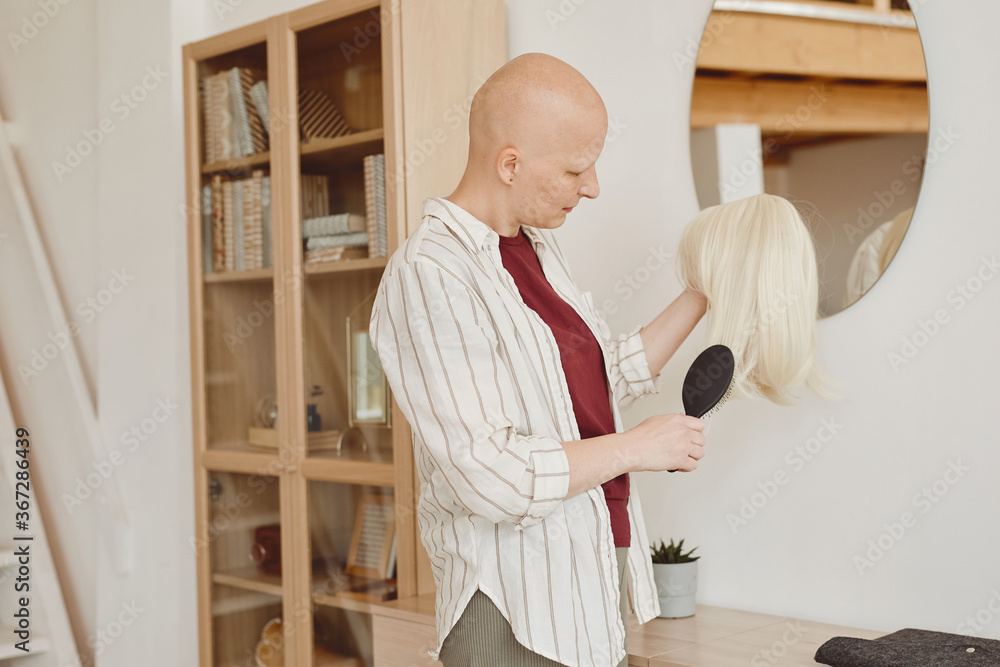 Warm-toned portrait of bald adult woman brushing wig while standing by mirror in modern home interior, alopecia and cancer awareness, copy space - obrazy, fototapety, plakaty 