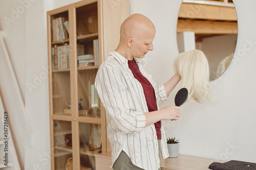 Warm-toned portrait of bald adult woman brushing wig while standing by mirror in modern home interior, alopecia and cancer awareness, copy space