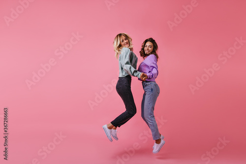 Debonair shapely girls holding hands and smiling. Studio portrait of jumping female friends expressing happiness.