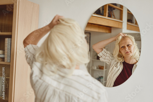 Fotografie Warm-toned back view portrait of bald woman taking off wig while looking in mirr