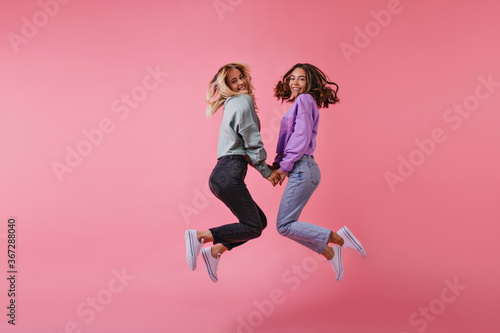 Fototapeta Naklejka Na Ścianę i Meble -  Studio portrait of blithesome best friends holding hands on pink background. Charming sisters in trendy pants jumping and laughing.