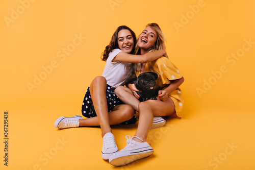 Stunning brunette girl in white shoes embracing her sister with happy smile. Carefree blonde lady having fun with best friend and bulldog during photoshoot on yellow background.