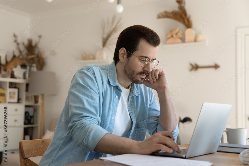 Focused young Caucasian man in glasses sit at desk at home look at laptop screen browsing surfing web, pensive millennial male work on computer, consider paperwork using gadget, technology concept Focused young Caucasian man in glasses sit at desk at home look at laptop screen browsing surfing web, pensive millennial male work on computer, consider paperwork using gadget, technology concept