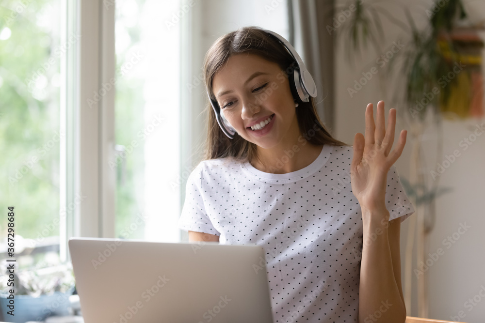 Smiling millennial girl in earphones wave greeting talk speak on video call on laptop at home, happy young woman in headphones have webcam virtual conversation or communication on modern computer Smiling millennial girl in earphones wave greeting talk speak on video call on laptop at home, happy young woman in headphones have webcam virtual conversation or communication on modern computer