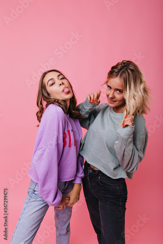 Spectacular girl in purple shirt posing with tongue out. Indoor photo of confident blonde lady fooling around with best friend.