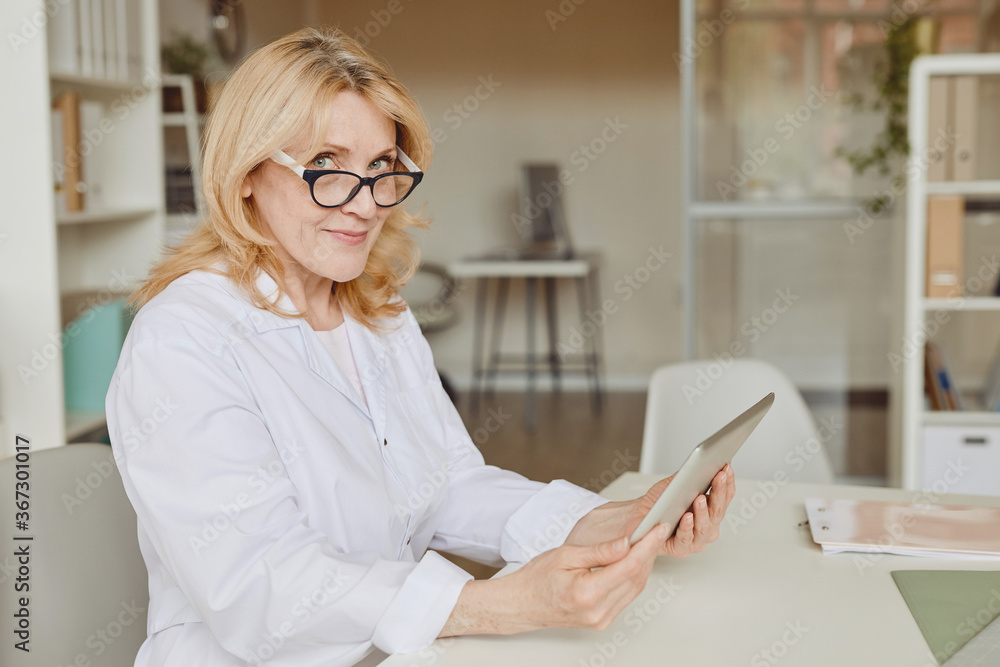Warm-toned waist up portrait of mature female smiling at camera and holding tablet while working in modern clinic, copy space Warm-toned waist up portrait of mature female smiling at camera and holding tablet while working in modern clinic, copy space