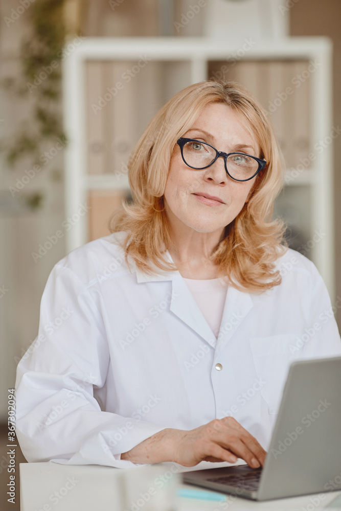 Vertical warm-toned portrait of mature female doctor looking at camera while working at desk in modern clinic office Vertical warm-toned portrait of mature female doctor looking at camera while working at desk in modern clinic office
