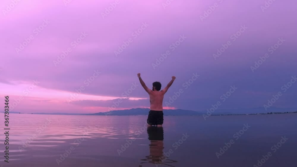 Man in swimming trunks in the water in a lake with his arms raised during a sunset Man in swimming trunks in the water in a lake with his arms raised during a sunset