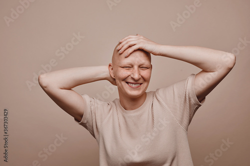 Warm-toned waist up portrait of carefree bald woman touching shaved head and smiling while posing against minimal beige background in studio, alopecia and cancer awareness, copy space
