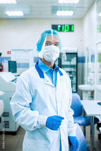 A medical worker, a laboratory assistant in a protective mask, visor, disposable cap and rubber gloves stands in a laboratory, hospital.