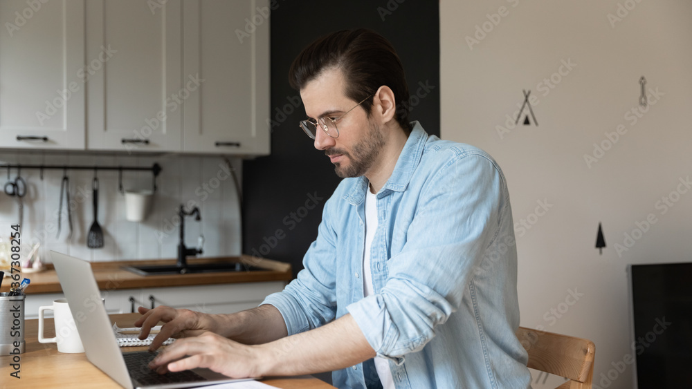 Concentrated young Caucasian man in glasses look at laptop screen browsing surfing web on laptop at home, focused millennial male freelancer work distant online on computer, sitting at kitchen counter