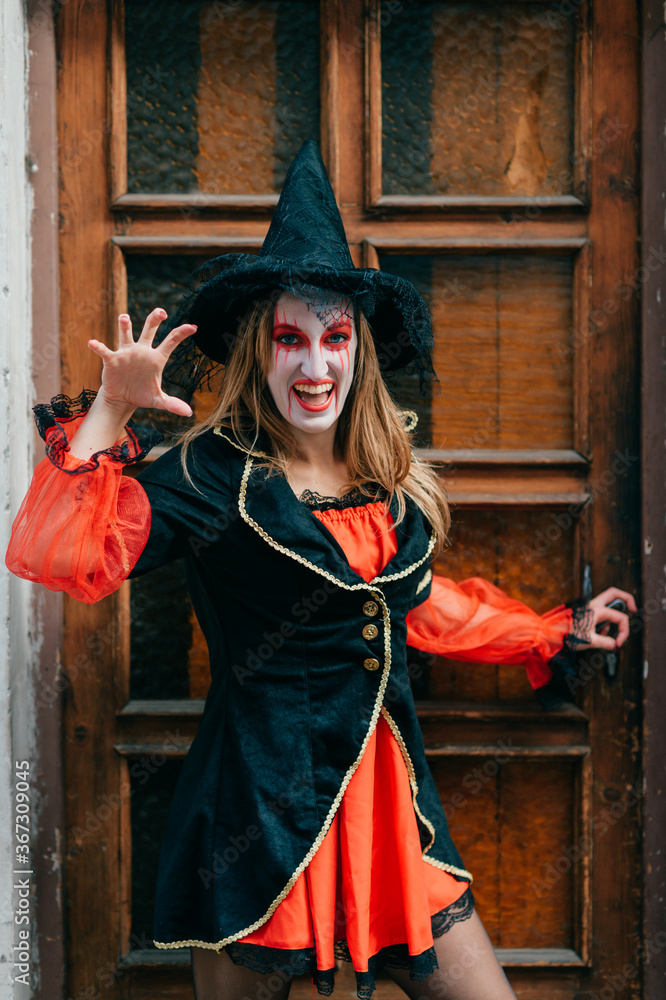 Charming female in witch costume poses outside in front of the big old door in black and red dress