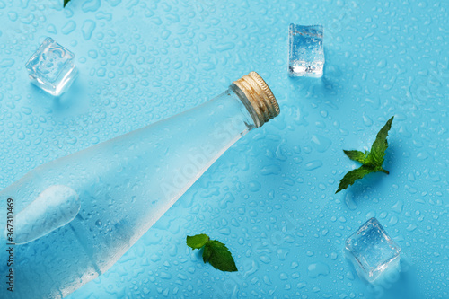 Bottle with an ice cold beverage, ice cubes, drops and mint leaves on a blue background.