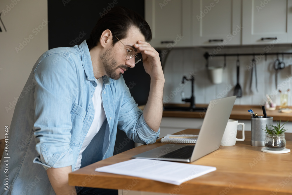 Troubled young Caucasian man sit in kitchen look at laptop screen thinking pondering of problem solution, pensive male freelancer work on computer, make decision studying or analyzing email on gadget - obrazy, fototapety, plakaty 