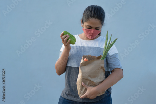 Young Indian girl wearing hand stitched cotton mask looking into grocery bag with vegetables. Budget buying the necessities during lockdown. Grey background with copy space photo