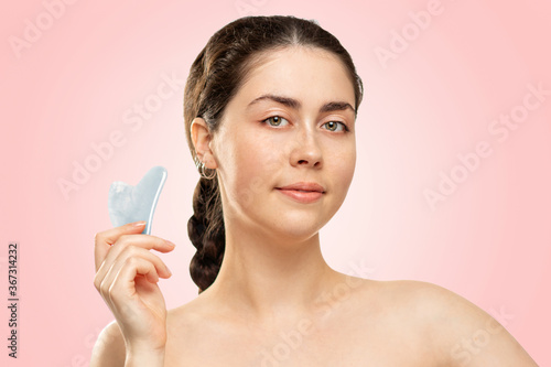 Gua Sha Massage. A portrait of a woman in a white bath towel holds a quartz gua Sha scraper in her hand. Pink background. Copy space. Concept of alternative medicine and cosmetology