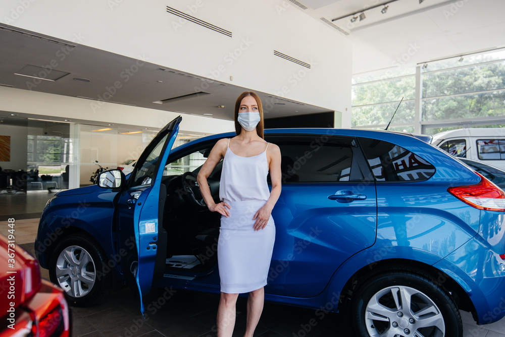 A young pretty girl inspects a new car at a car dealership in a mask during the pandemic. The sale and purchase of cars, in the period of pandemia