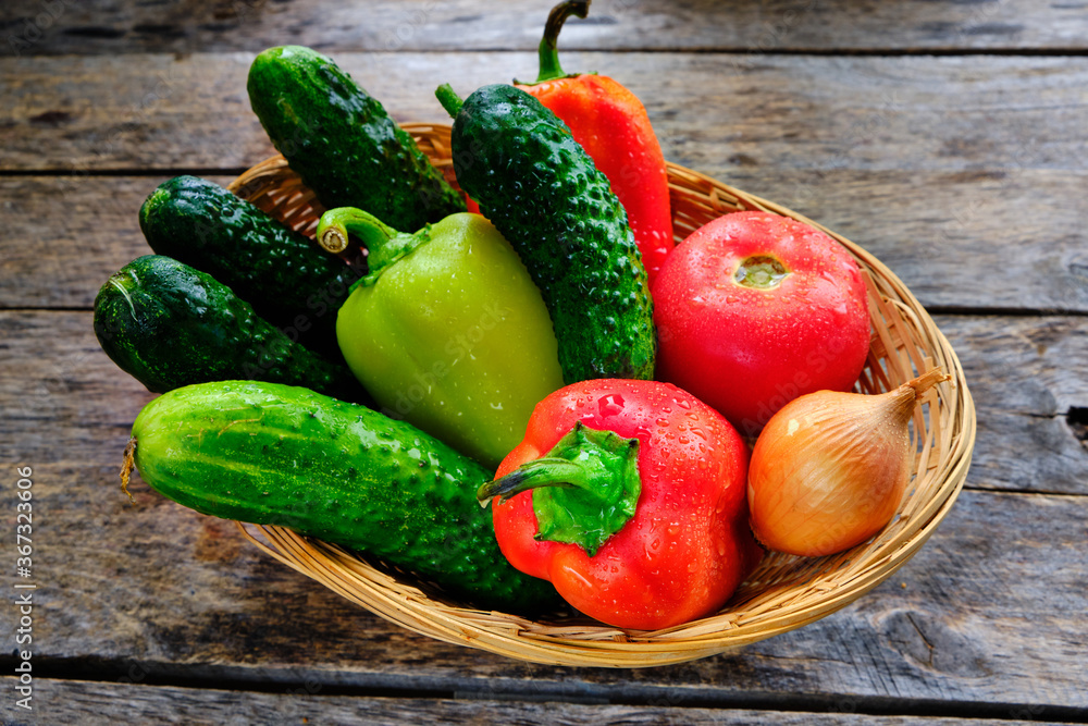 Cucumbers, tomatoes, sweet bell peppers and onions in a basket on a wooden table in a rustic style.
