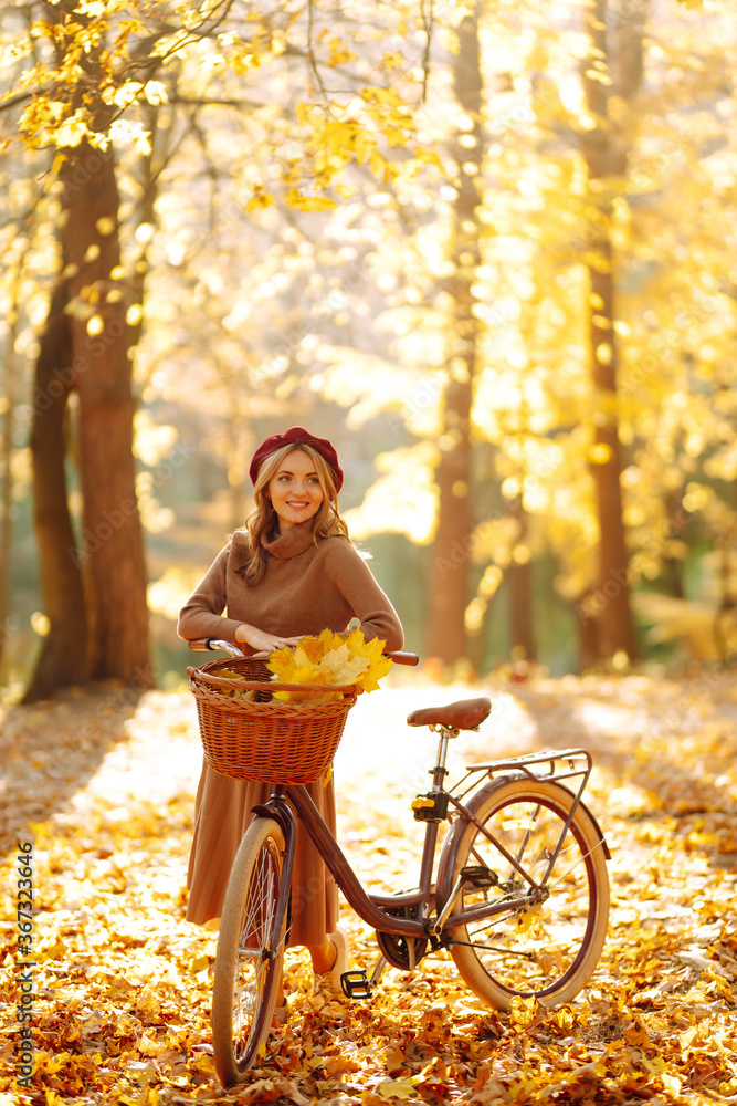 Young smiling girl with a bicycle walks in the autumn forest at sunset. - obrazy, fototapety, plakaty 