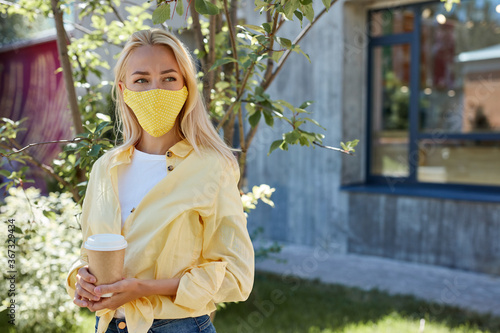 attractive blonde female in medical mask looks away, hold cup of coffee in hands, walk in the park after quarantine photo